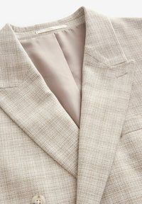 Beige blazer featuring a textured woven pattern, notch lapels, and a smooth tan lining. Detailed with a single button closure.