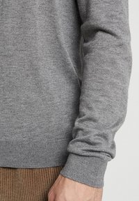 Grå stickad tröja med mjuk textur, åtsittande ärmavslut och rak fåll. Tyget verkar lätt och andningsbart.