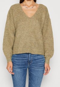 Maglione in maglia khaki con scollatura a V, texture a coste e maniche a palloncino, abbinato a jeans in denim blu. I dettagli dei punti sono visibili.