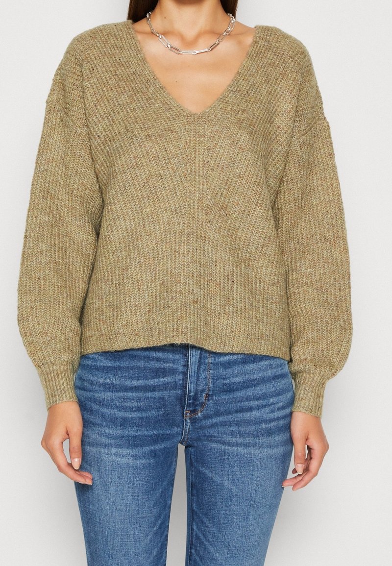 Maglione in maglia khaki con scollatura a V, texture a coste e maniche a palloncino, abbinato a jeans in denim blu. I dettagli dei punti sono visibili.
