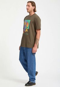 Olivgrünes Baumwoll-T-Shirt mit buntem Grafikdesign von Sonnen und einer Katze, kombiniert mit lockeren blauen Jeans und schwarzen Schuhen.