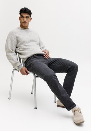 Jeune homme portant un pull gris, un jean noir et des chaussures beiges, assis de manière détendue sur une chaise en métal, sur un fond blanc uni.