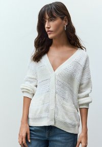 Cardigan tricoté blanc avec un décolleté en V profond, présentant des motifs texturés, des manches retroussées et des fermetures à boutons, associé à un jean bleu.