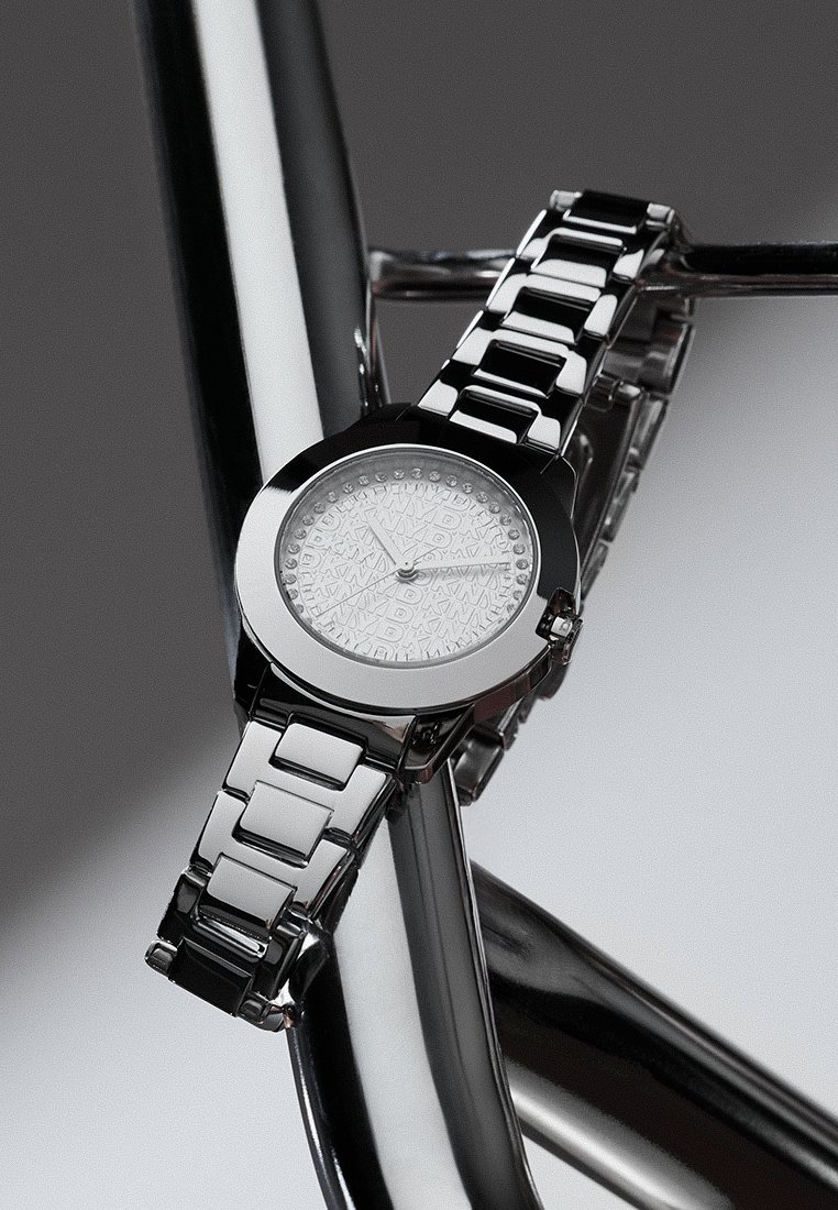 Reloj de pulsera de metal plateado con una pulsera de eslabones, cara redonda con un dial decorado y pequeños cristales marcando las horas, descansando sobre barras metálicas reflectantes.