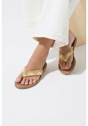 Piedi con infradito marroni con cinturini decorati con perline gialle e beige, abbinati a pantaloni bianchi a gamba larga, in piedi su una superficie chiara.