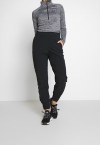 Šedé tričko s dlouhým rukávem a zipovým límcem v kombinaci s černými tepláky s elastickým pasem, postranními kapsami a manžetami na kotnících. Outfit doplňují sportovní boty.