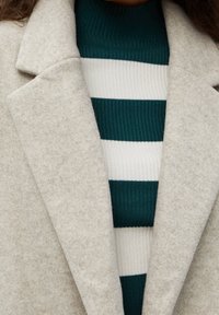 Manteau beige en mélange de laine avec un col cranté, superposé à un col roulé côtelé à rayures bleu sarcelle et blanc. Texture douce, design simple.