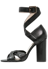 Mustat nahkakengät, joissa on korkea korko ja nilkkaremmi, joka kiinnitetään neliönmuotoisella kullanvärisellä soljella. Kengissä on kiertyvät etuvarpaat, jotka paljastavat avonaisen varpaankärjen.