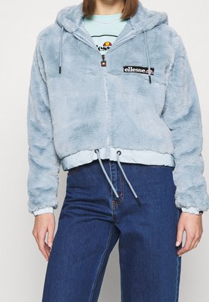 Personne portant une veste à capuche zippée duveteuse bleu clair avec le logo « ellesse », associée à un jean bleu taille haute et un t-shirt assorti.