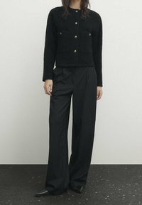 Svart texturerad jacka med guldknappar och framfickor, i kombination med vida svarta byxor med smalrandigt mönster och spetsiga svarta skor.