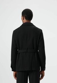 Zwarte gestructureerde jas met een kraag, een taille met ceintuur en knoopmanchetten, voorzien van een diagonaal geribd patroon en een getailleerde pasvorm.