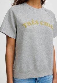 Grå kortärmad sweatshirt med ribbad halsringning och nederkant. Har guldtexten "TRÈS CHIC" över framsidan. Mjuk, texturerad tyg.