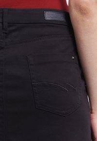 Schwarze Denim-Jeans mit einer Gesäßtasche, die ein gewundenes Nahtdesign und ein Logo-Tag aufweist. Glatte Textur, schmaler Schnitt und kontrastierende Nähdetails.