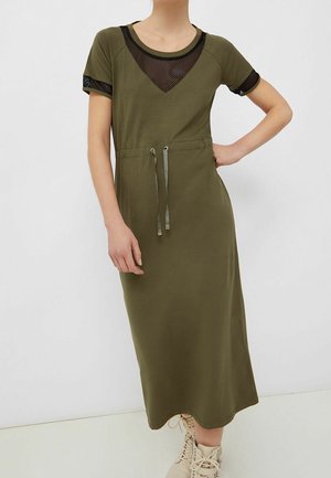 Persoon draagt een olijfgroene midi-jurk met zwarte mesh details bij de halslijn en mouwen, een aantrekkoord in de taille en beige veterlaarzen.