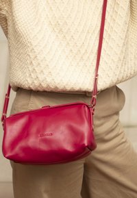 Borsa a tracolla rossa in pelle con texture liscia, chiusura a zip e una sottile tracolla, indossata in vita su un outfit chiaro.