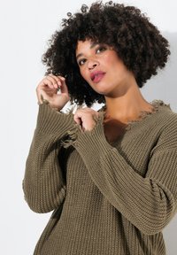 Pull en tricot vert olive texturé avec un col en V, des poignets effilochés et un ourlet effiloché. Le matériau est épais, avec une coupe décontractée et un motif à côtes distinct.
