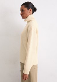 Roomkleurige, geribbelde coltrui met lange mouwen en een losse pasvorm. Zachte, gestructureerde stof met horizontale ribbels en een zoom met een split aan de zijkant.