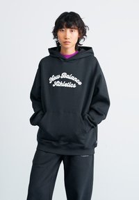 Jeune personne portant un hoodie noir "New Balance Athletics" et un pantalon noir assorti, se tenant les mains dans les poches devant un fond uni.