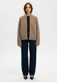 Cardigan beige à côtes avec un col montant et une fermeture éclair sur le devant, porté sur un top noir, associé à un jean en denim foncé à jambes larges et des escarpins noirs.