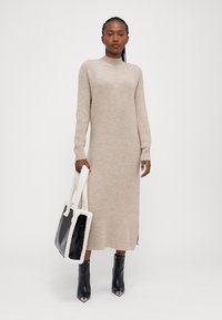 Πλεκτό μπεζ μακρύ φόρεμα με ψηλό λαιμό, ριγωτή υφή και πλάγιες σχισμές, συνδυασμένο με μια μαύρη και λευκή τσάντα tote και γυαλιστερές μαύρες ankle boots.