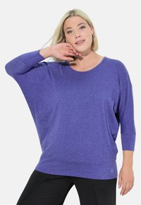 Paarse, loszittende top met driekwartmouwen, ronde halslijn, zachte textuur en een klein logo detail aan de zoom. Zwarte broek zichtbaar.