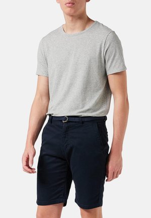 Mann trägt ein hellgraues Kurzarm-T-Shirt und schwarze Shorts mit einem gewebten Gürtel vor einem einfarbigen Hintergrund.
