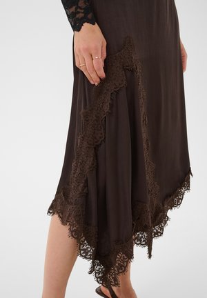 Personne portant une jupe marron avec une bordure en dentelle, debout. Une main repose sur la jupe, portant de fines bagues en or.