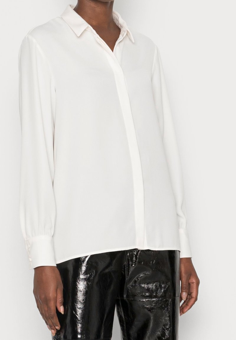 Camicia bianca a maniche lunghe con colletto a camicia, realizzata in tessuto liscio, con orlo dritto e dettagli in bottoni sui polsini.
