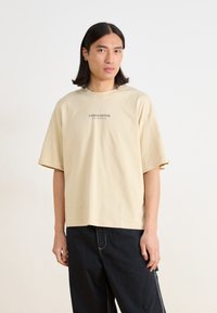 Beige oversized bomull t-skjorte med korte ermer, med en liten "STUDIO EDITION" trykk i midten. Brukt med svarte bukser.