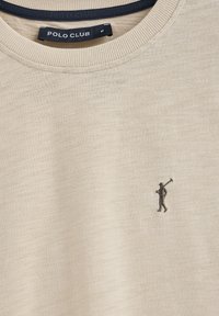 T-shirt beige a maniche corte con scollatura a costine, che presenta un piccolo figura ricamata scura che tiene una mazza da golf sul davanti.
