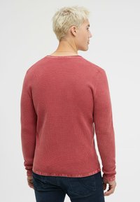 Rotes Langarmshirt mit einem strukturierten Waffelstrickmuster. Verfügt über einen Rundhalsausschnitt und kontrastierende Nähte entlang des Saums und der Bündchen.