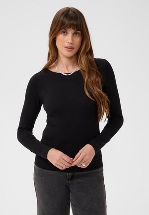 Femme aux longs cheveux bruns portant un haut noir à manches longues, un jean gris, un collier en argent et une bague, debout devant un fond uni.