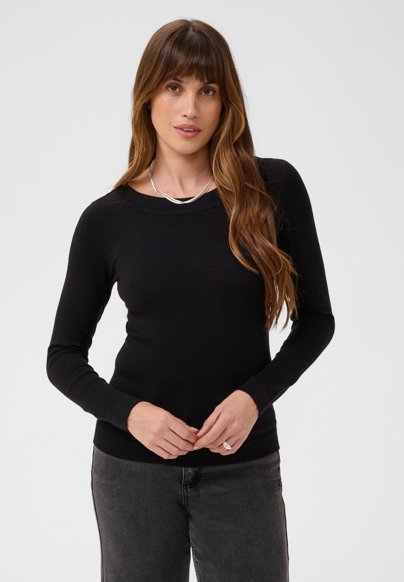 Femme aux longs cheveux bruns portant un haut noir à manches longues, un jean gris, un collier en argent et une bague, debout devant un fond uni.