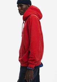 Červená mikina s kapucňou na šnúrku, predným vreckom a tónovým vzorom na rukáve. Nosená cez karovanú košeľu, z ktorých jedna má voľný strih a mäkkú textúru.