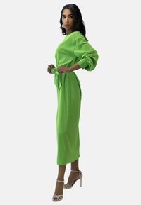 Groene lange mouwen jurk met een ceintuur in de taille en zijzakken, gemaakt van gladde stof. Het model draagt doorzichtige sandalen met hoge hak.