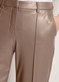 Close-up van taupe leren broeken met riemlussen, details van de voorknaad en een hand gestoken in de rechterzak.