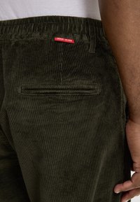 Pantalon en velours côtelé vert olive avec une texture côtelée, comportant une poche arrière et une étiquette rouge "JOOP JEANS" à la taille.