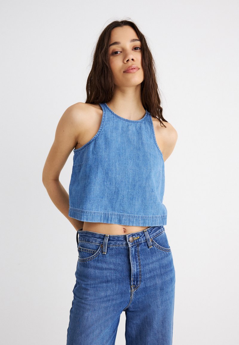 Ung kvinde med langt mørkt hår iført ærmeløs blå denim crop top og højtsiddende blå jeans, stående mod en ensfarvet hvid baggrund.
