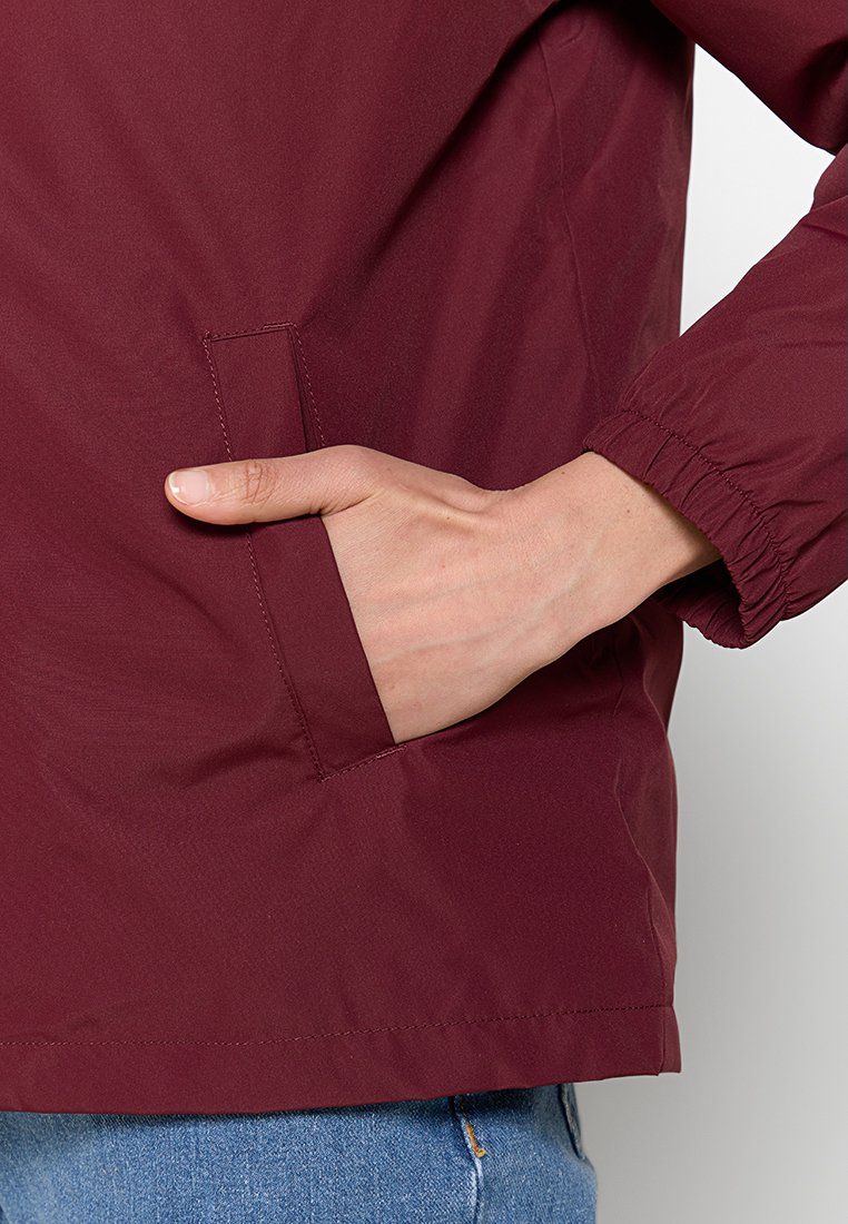 Chaqueta ligera color burdeos con una textura suave, puños elásticos y un bolsillo lateral. Primer plano de una mano descansando sobre el bolsillo.