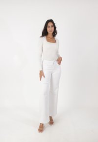 Witte wijde jeanstof en een aansluitende beige top met lange mouwen en een vierkante halslijn. Het model staat met één hand op haar heup, met doorzichtige sandalen met hakken.