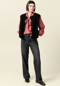 Svart fleeceväst över en röd, plisserad blus med en slipskrage. Modellen bär grå vida jeans och svarta loafers. Enkelt bakgrund.