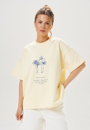 Młoda kobieta z jasnymi włosami w koku, ubrana w bladożółtą, oversize'ową koszulkę z grafiką palm i tekstem o klubie sportowym, zdrowotnym i wellness.