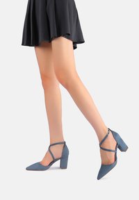 Scarpe blu scamosciate con tacco a blocco e punta appuntita, con cinturini incrociati alla caviglia, abbinate a una gonna nera, che mette in risalto una texture liscia.