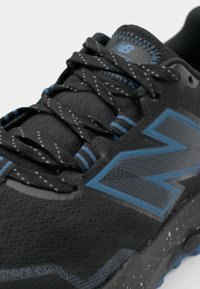 Chaussure de sport noire avec surface texturée, accents bleus et lacets à motifs. Présente un logo "N" bien en évidence et un design de semelle mouchetée.