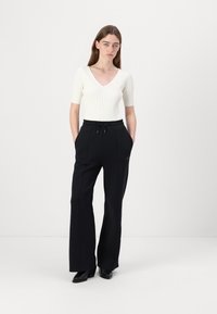 Schwarze weite Hosen mit einem Zugbandbund, kombiniert mit einem taillierten weißen gerippten V-Ausschnitt-Oberteil. Glatte Textur, minimalistisches Design, keine sichtbaren Muster.