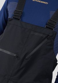 Czarne wodoodporne ogrodniczki z regulowanymi paskami, kieszenią na zamek z przodu i subtelnym logo. Nosi się je na niebieskiej koszuli z długim rękawem w paski.