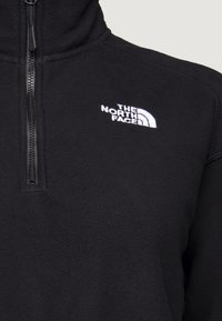 Pull polaire noir avec fermeture éclair quart de zip et logo The North Face brodé en blanc sur la poitrine gauche.