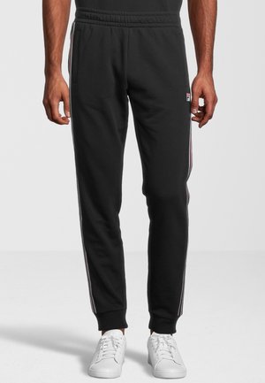 Homme portant un pantalon de jogging noir avec des bandes latérales et des baskets blanches, debout, les bras détendus le long du corps, devant un fond uni.
