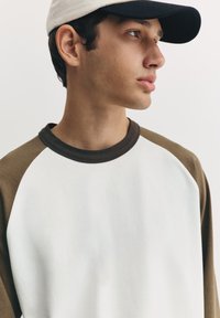 Bluză cu mâneci raglan având corpul alb și mânecile verde măsliniu, guler rotund în maro închis și o șapcă de baseball deschisă la culoare.