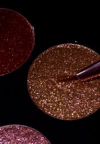 Two circular glitter pigments: one deep red and one golden bronze. Both feature a fine, shiny texture, with a brush partially immersed in the bronze.